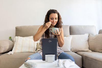 Content female in domestic outfit sitting on sofa with cup of coffee and watching movie on tablet while relaxing at weekend at home