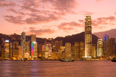 Illuminated buildings by river against sky during sunset