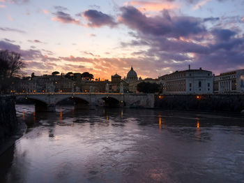 Bridge over river by buildings against sky at sunset