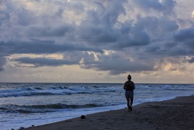 Rear view of woman walking at beach against cloudy sky during sunset