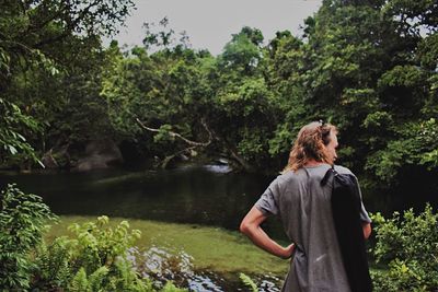 Rear view of young man with hand on hip standing against lake in forest