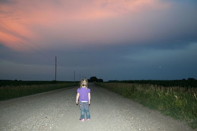 Rear view full length of girl standing on road amidst grassy field against sky during sunset