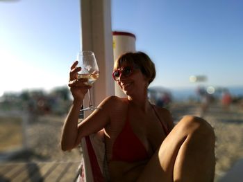 Happy woman in bikini having wine while sitting on chair at beach