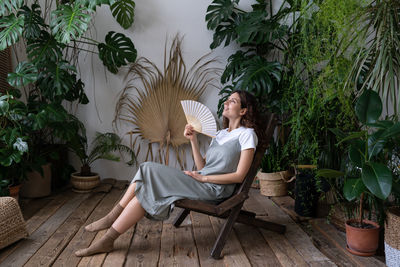 Happy woman florist refreshing in home garden breathing fresh air hold paper fan in hand at hot day