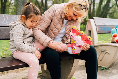 Positive mature woman with blond hair in warm casual clothes helping cute granddaughter to put on roller skates sitting on wooden bench in park