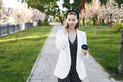 Young woman holding coffee talking over smart phone while standing on footpath