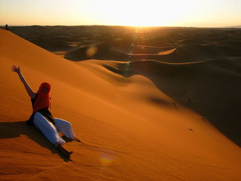 Smiling young woman with arm raised sitting on sand dune during sunny day