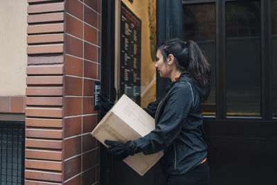 Female messenger ringing intercom while carrying box by closed door
