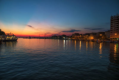 Illuminated buildings by river against sky at sunset