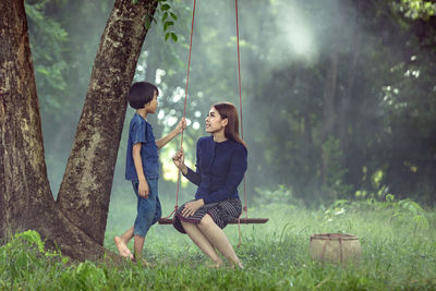 Girl by young woman sitting on rope swing in forest