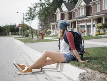 Side view of woman with backpack sitting on footpath against houses