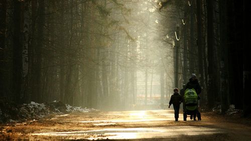 Woman pushing baby stroller while walking with boy on road amidst trees during winter