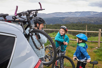 Side view of positive mother with kids in helmets taking bicycle from trunk rack of modern car parked in countryside