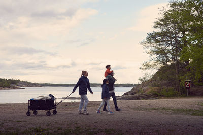 Woman pulling camping cart while walking with family at beach against sky