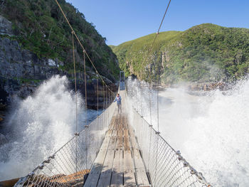 Sea waves splashing on woman standing at rope bridge