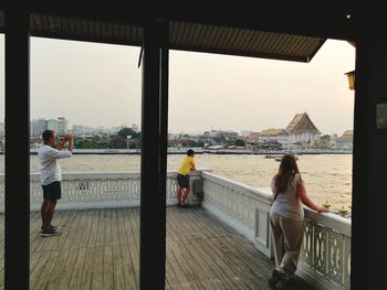 People standing on railing by sea against sky in city