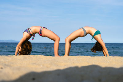 Side view of friends wearing bikinis exercising at beach against sky
