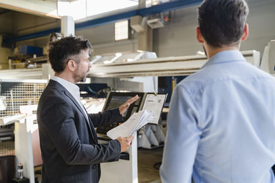 Businessman with paper having discussion with colleague while standing against machine at factory