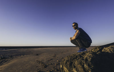 Side view of young man crouching on rock against clear sky