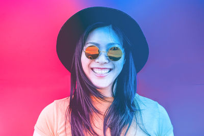 Portrait of smiling young woman wearing sunglasses and hat against colored background