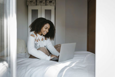 Smiling woman using laptop while sitting on bed at home