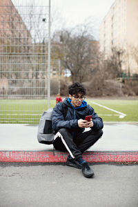 Full length of young man using mobile phone while sitting on sidewalk against soccer field during winter