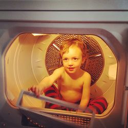 Playful boy sitting in washing machine seen through glass at home