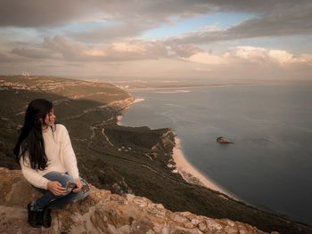 Woman sitting on cliff against sea during sunset