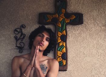 Portrait of young man with hands clasped standing by cross on wall at home