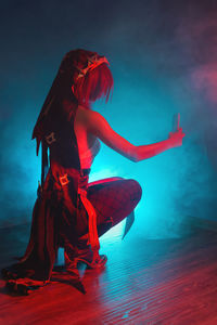 Vertical photo of a young girl in a cosplay costume sitting in a room with colored smoke and light