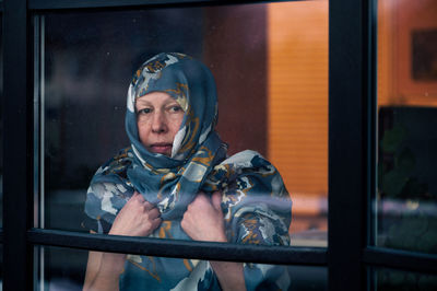 Sad middle-aged woman with scarf on head looking through the dirty window glass