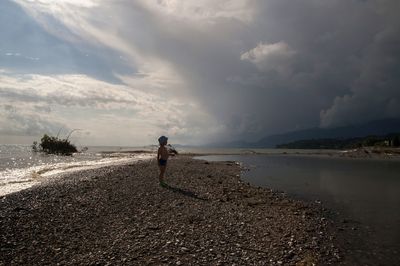 Shirtless boy standing at beach against cloudy sky