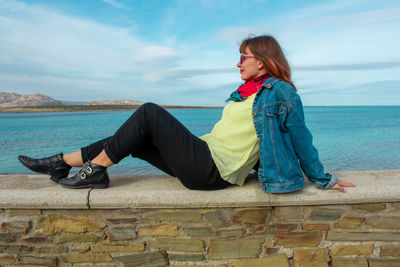 Man sitting on retaining wall by sea against sky