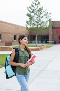 Young student in a backpack holding books standing in front of a school 