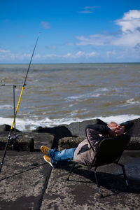 Rear view of man sitting on chair by fishing rod at rocky sea shore