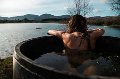 Anonymous woman chilling in barrel with hot water and algae and observing calm lake on weekend day in countryside