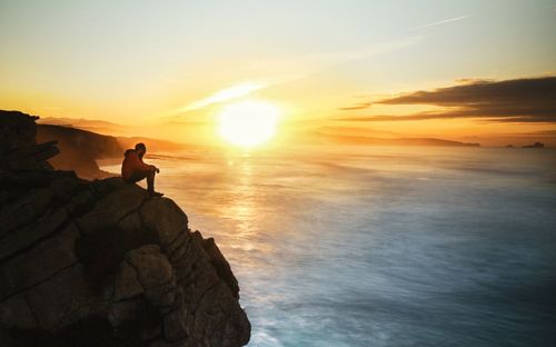 Man sitting on rocks by sea against sky during sunset
