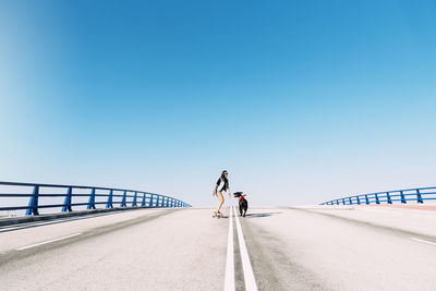 Dog running with woman skateboarding against blue sky