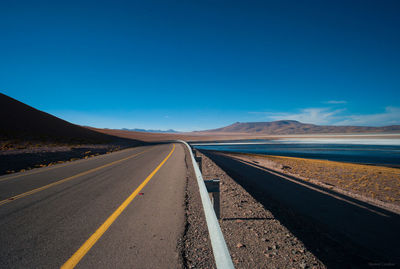 Empty road by lake against clear blue sky on sunny day