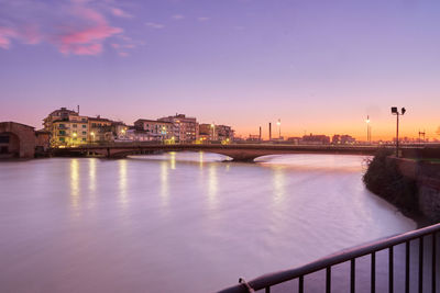 Illuminated bridge over river by buildings against sky at sunset