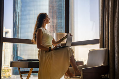 Side view of young woman using digital tablet while sitting on window