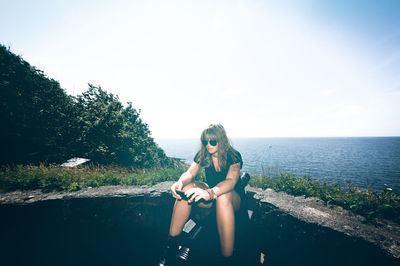 Young woman using mobile phone while sitting on retaining wall against sea