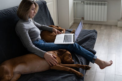 Woman sitting at couch with twovizsla dog, stroking, remote working on laptop. love pets. side view.