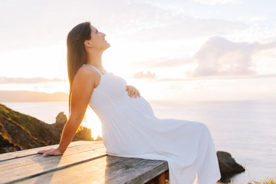 Pregnant woman sitting on bench against sea