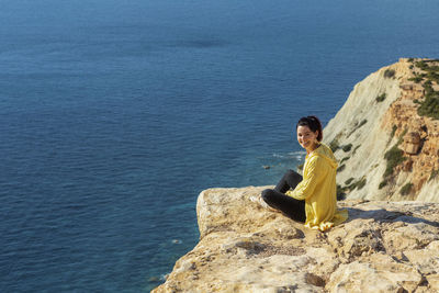 Young woman in yellow zip jacket sitting on a cliff turning back to the camera