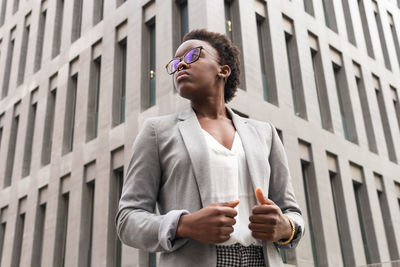 Low angle of young female entrepreneur in classy jacket and eyeglasses standing near modern skyscrapers in city