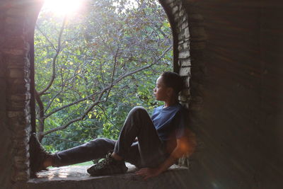 Thoughtful boy sitting on window sill against branches