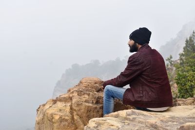 Rear view of man sitting on rock against sky