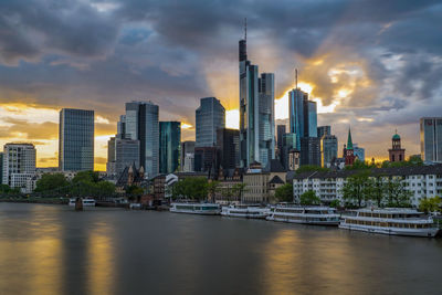 Modern buildings by river against sky during sunset