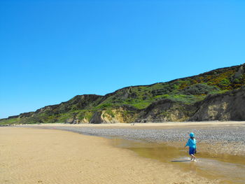 Rear view of teenage girl walking at beach against clear blue sky